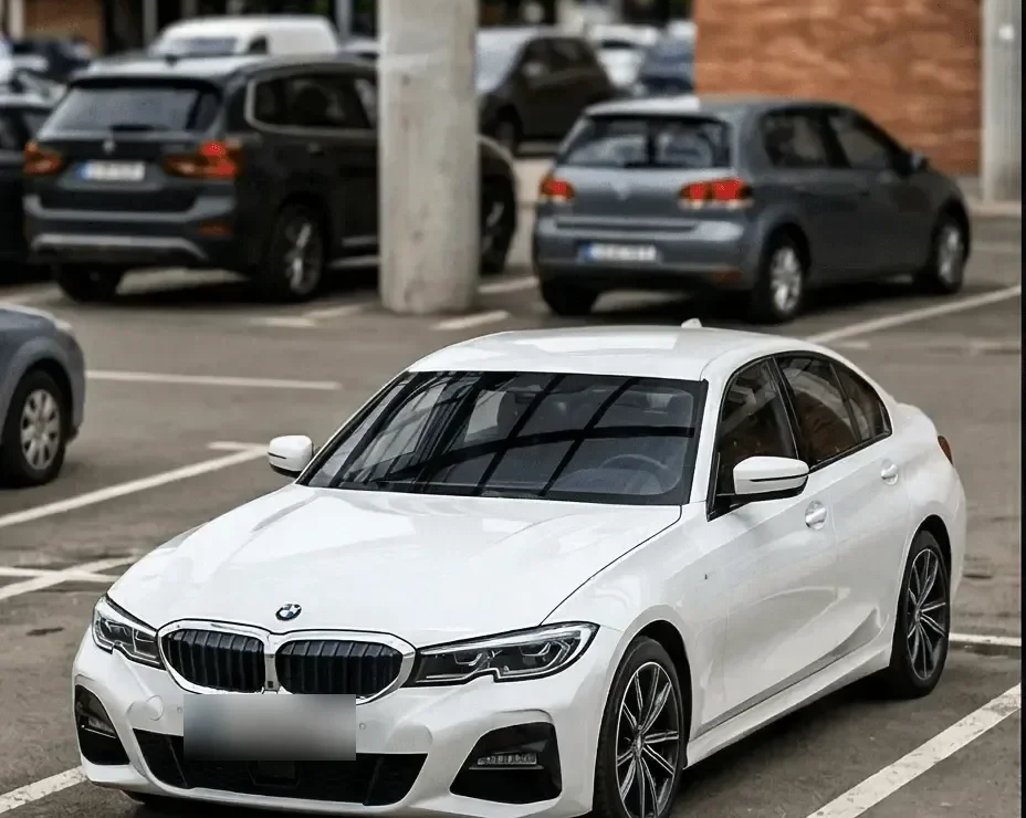 A white BMW 3 Series parked in an outdoor lot beside other cars and a concrete pillar