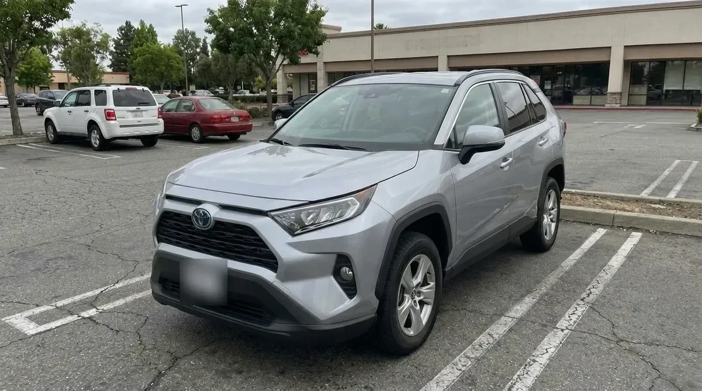 A silver Toyota RAV4 parked on an open lot on an overcast day, with other parked cars, trees and a low commercial building behind it